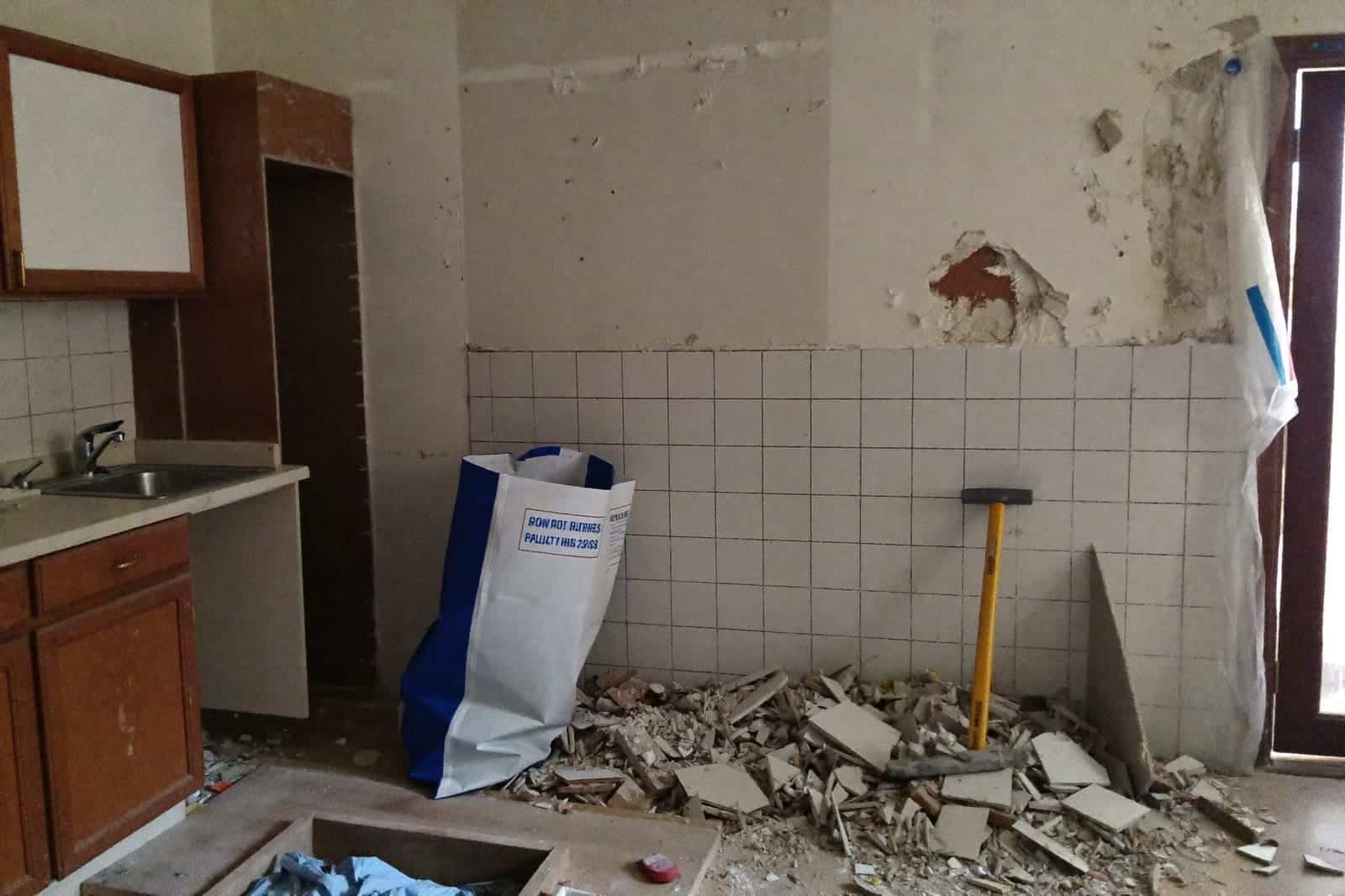 Half uitgebroken oude keuken met puin op de vloer, afgeslagen tegels en gereedschap tegen de muur